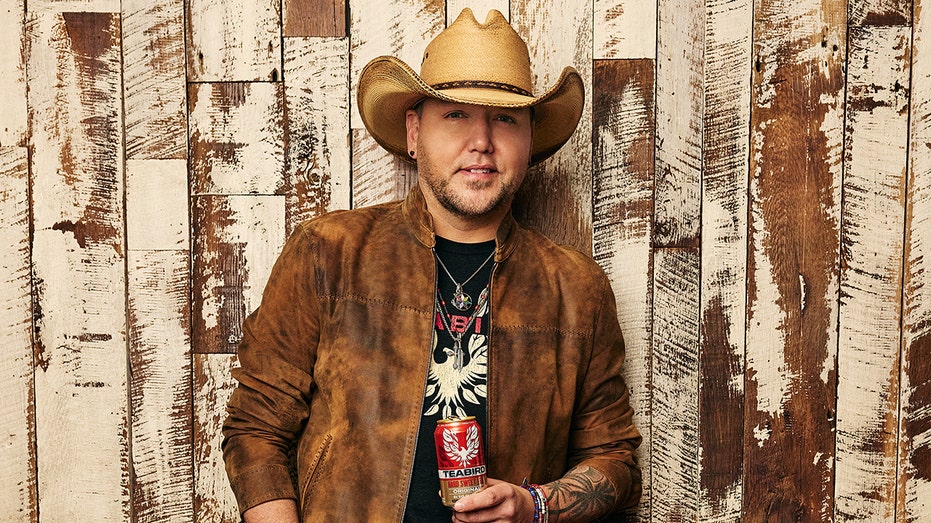 Jason Aldean in a brown coat and tan cowboy hat holding his Teabird