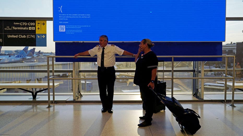 United Airlines employees wait by a departures monitor displaying a blue error screen, also known as the 