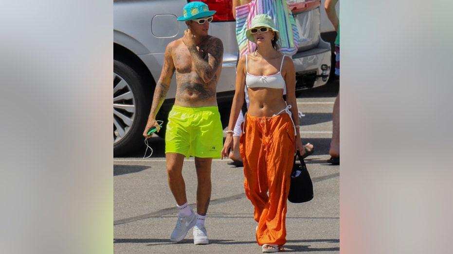 Justin Bieber in neon yellow swim trunks and a turquoise bucket hat walks alongside Haiely Bieber in a white bikini top and orange pants