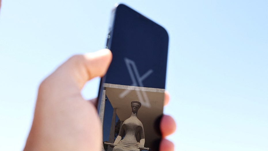 The logo of social media platform X is seen on a mobile phone next to a reflection of Alfredo Ceschiatti's sculpture The Justice, outside Brazil's Supreme Court in this illustration taken August 30, 2024, in Brasilia