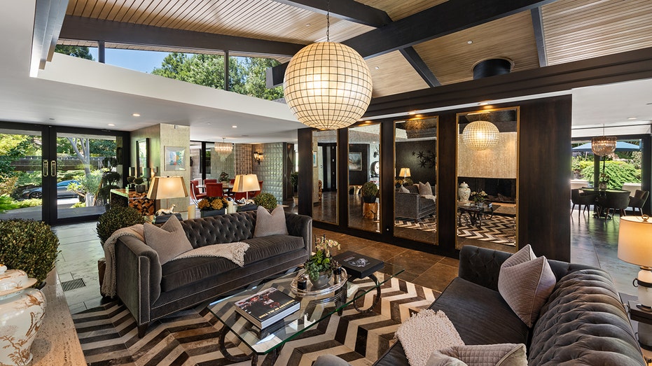 The living room boasts high wood-paneled ceilings, brown tiled floors and a wall lined with mirrors.