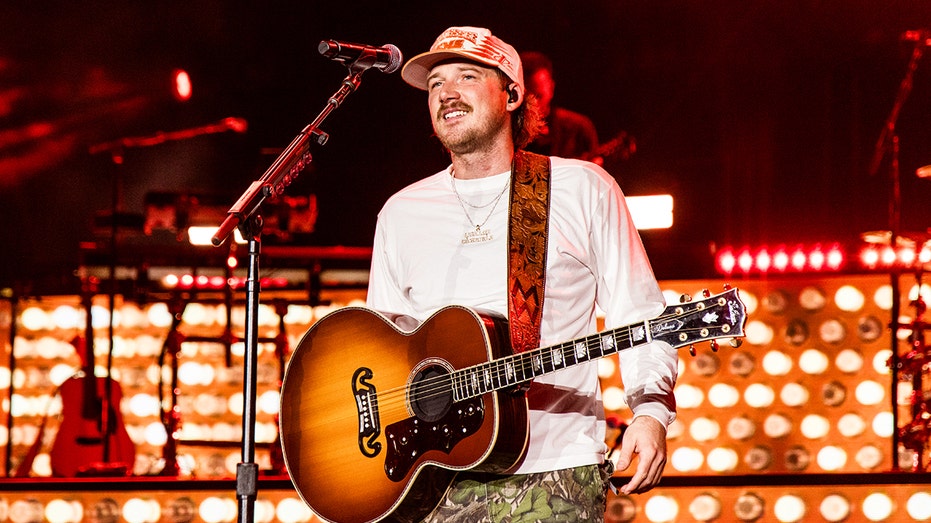 Country star Morgan Wallen smiles wearing a guitar on stage.