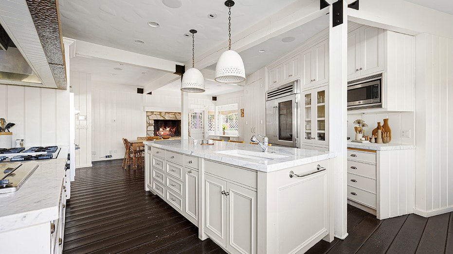 The all-white kitchen features a large center island and a fireplace next to a breakfast nook.