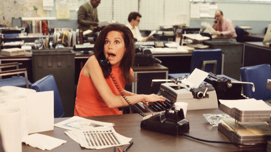 Aktris Amerika Mary Tyler Moore mengucapkan kejutan di telepon sambil mengetik saat orang lain bekerja di latar belakang dalam sebuah adegan dari 'The Mary Tyler Moore Show' (juga dikenal sebagai 'Mary Tyler Moore'), Los Angeles, California, awal tahun 1970-an. Moore mengenakan gaun oranye tanpa lengan saat dia duduk di belakang mejanya. (Foto oleh Arsip Foto CBS/Getty Images)