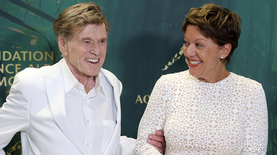 Robert Redford in a white button down and blazer holding onto the arm of his wife Sibylle Szaggars in a sparkling white dress