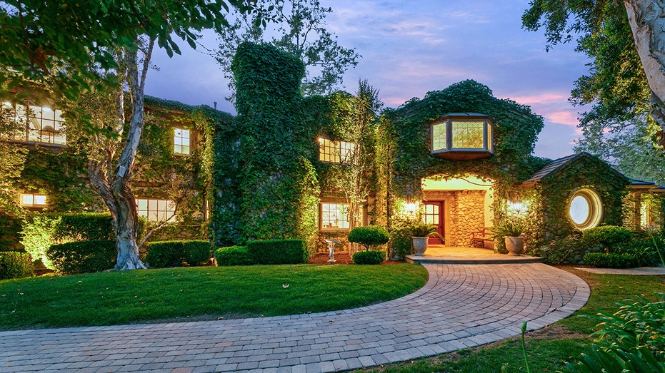 A lengthy driveway leads to an ivy-covered house surrounded by trees and grass.