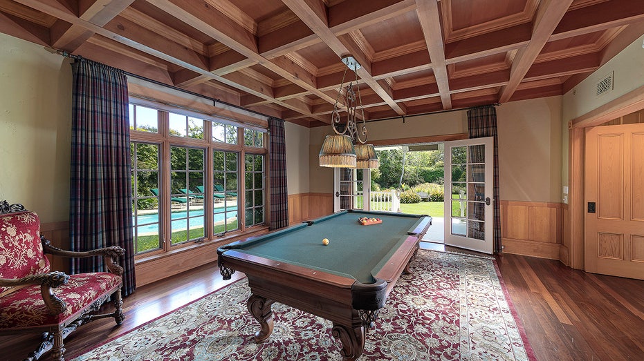 The billiards room features coffered ceilings and views of the swimming pool outside.