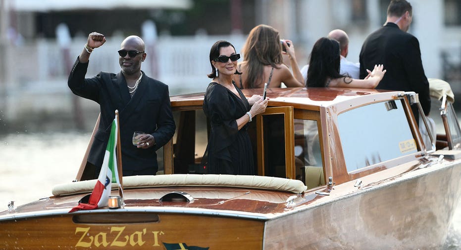 US manager Corey Gamble and US television personality Kris Jenner stand on a taxi boat as they leave the Gritti Palace Hotel ahead of the wedding of Amazon's founder Jeff Bezos with Lauren Sanchez in Venice on June 26, 2026. Celebrities in superyachts sail into Venice this week for the three-day wedding party of Amazon tycoon Jeff Bezos and Lauren Sanchez, despite irate locals who say the UNESCO city is no billionaire's playground. The tech magnate and journalist have reportedly invited about 200 guests to their multi-million dollar nuptials in the Italian city, which are expected to kick off on June 26 and end Saturday with a ceremony at a secret location. (Photo by Marco BERTORELLO / AFP) (Photo by MARCO BERTORELLO/AFP via Getty Images)