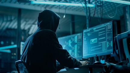 Shot from the Back to Hooded Hacker Breaking into Corporate Data Servers from His Underground Hideout. Place Has Dark Atmosphere, Multiple Displays, Cables Everywhere. - Fox Business News