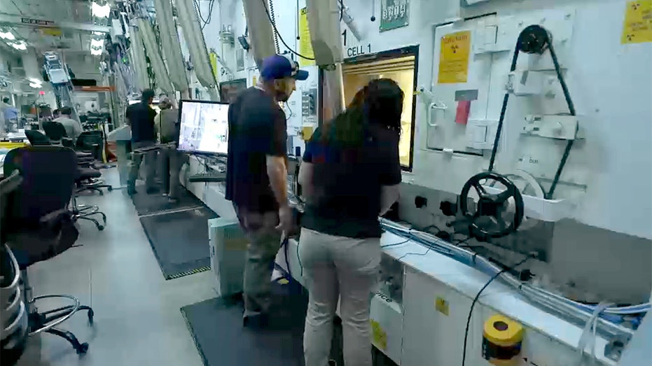 People in an industrial laboratory look into a window with an experiment inside.