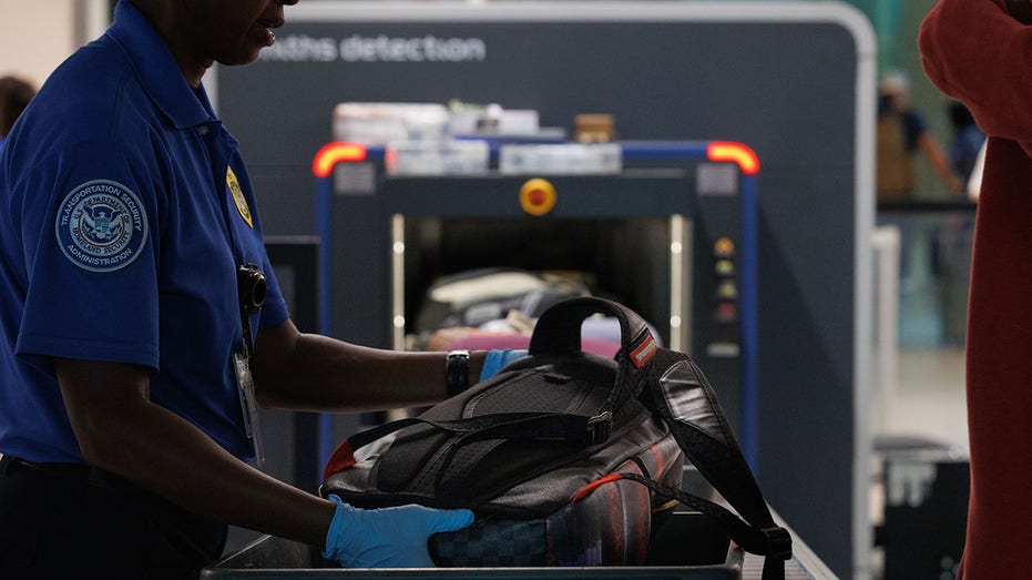 A Transportation Security Administration (TSA) agent assists travelers as they place their luggage onto a scanner belt.