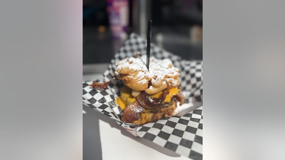 A grilled hamburger patty with crispy bacon and queso cheese sauce, sandwiched between two funnel cakes topped with powdered sugar.