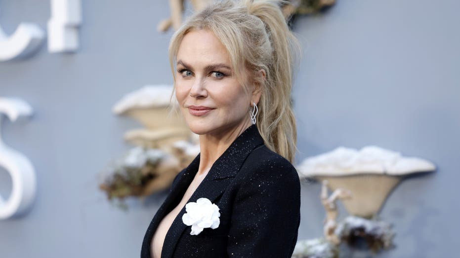 Nicole Kidman smiles, dressed in black blazer with a white flower that is fenced in lapel