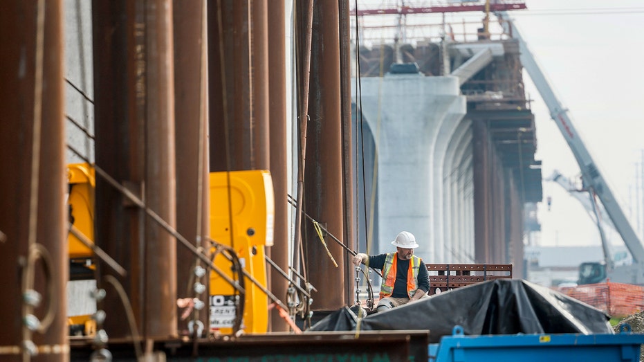 Heavy machinery and workers are visible on a long elevated concrete structure spanning farmland in California’s Central Valley.