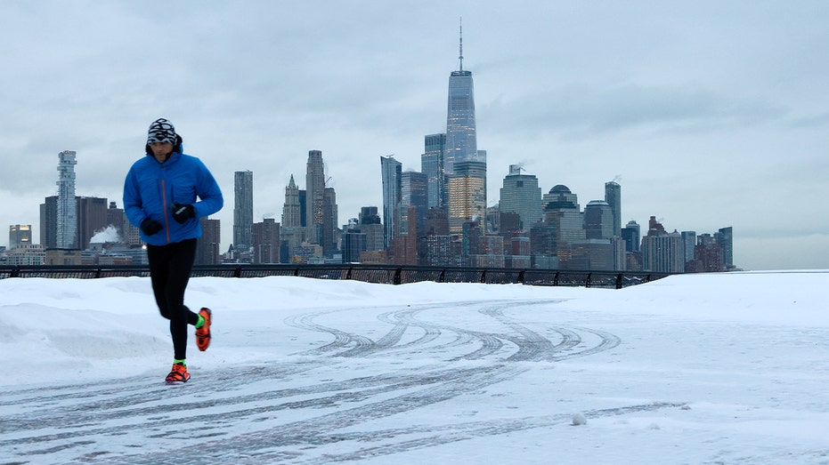 A jogger runs along a snowy waterfront path with the Manhattan skyline in the background.