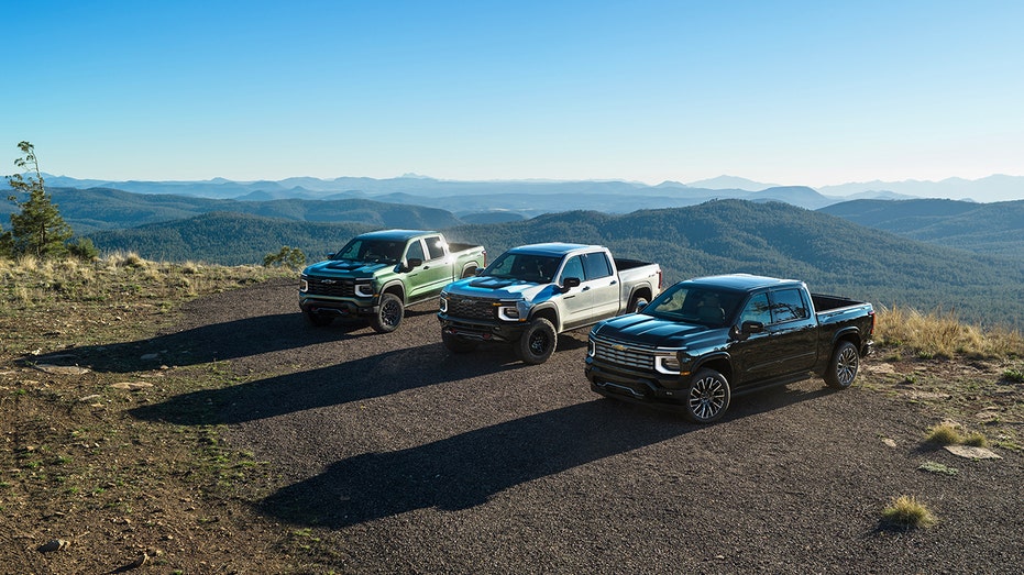 A 2027 Chevrolet Silverado 1500 Trail Boss in Trillium Metallic, a Silverado 1500 ZR2 in White Sands and a Silverado 1500 High Country in Black are shown, from left, on a mountain overlook.