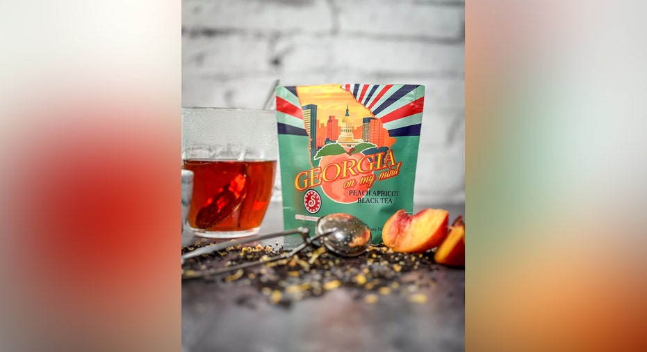A package of Georgia on My Mind peach-apricot black tea displayed beside a glass of tea, loose tea leaves and sliced peaches.