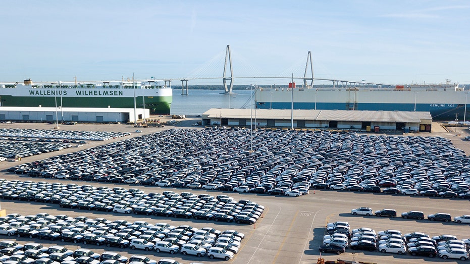 Vehicles await export at the Port of Charleston in Charleston, S.C., with cargo ships and the Arthur Ravenel Jr. Bridge in the background.