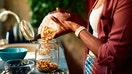 Close-up of a woman's hands pouring granola from a paper bag into a glass jar, with a bowl of blueberries in the foreground, suggesting a healthy breakfast preparation - Fox Business News