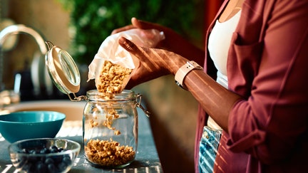Close-up of a woman's hands pouring granola from a paper bag into a glass jar, with a bowl of blueberries in the foreground, suggesting a healthy breakfast preparation - Fox Business News