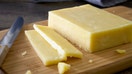 A block of cheddar cheese cut into slices on a chopping board. On a rustic wooden kitchen table kitchen utensils and herbs in the background. - Fox Business News