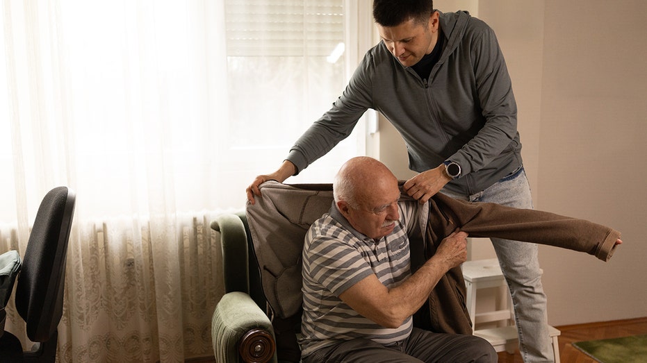 Caring middle aged son helping his senior father to get dressed while sitting in armchair at home