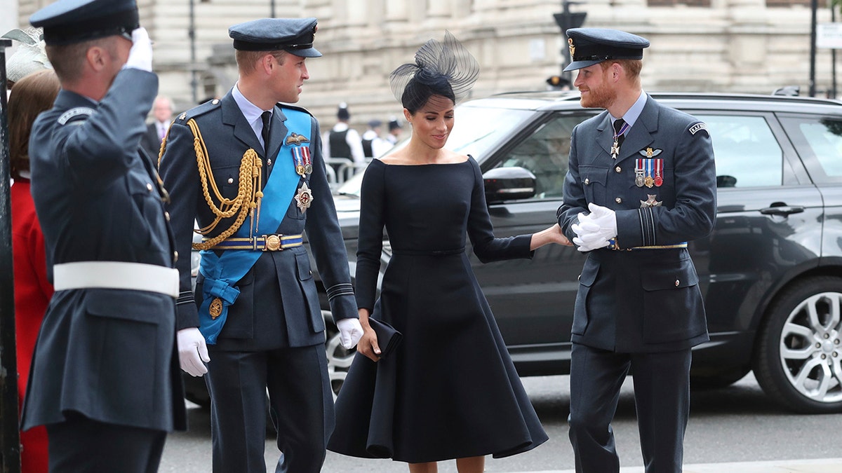 Meghan Markle wearing a black dress in between Prince William and Prince Harry as they wear military suits.