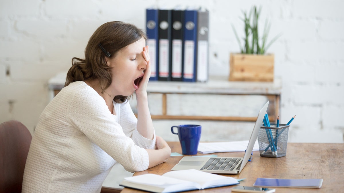 Portrait of young woman sitting at table in front of laptop, sleepy, tired, overworked, lazy to work. Attractive business woman yawning in home office relaxing or bored after work on laptop computer