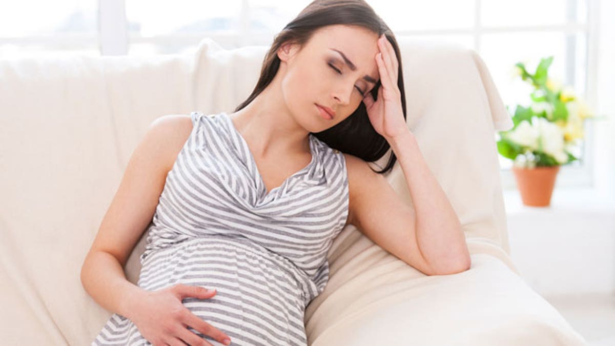 Struggling with morning sickness. Depressed pregnant woman holding hand on head and keeping eyes closed while sitting on a couch