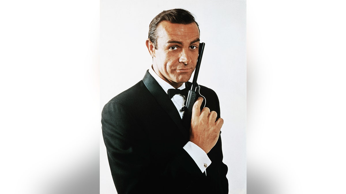 (Original Caption) Waist-up portrait of Sean Connery, as James Bond, caressing the barrel of a gun against the side of his face. Connery is wearing a tuxedo and bow tie and smiling slightly.