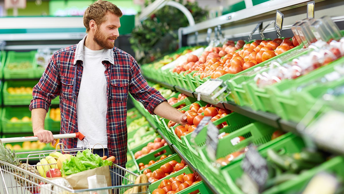 Hombre guapo con carrito de la compra tocando un tomate para definir su calidad en la sección de frutas y verduras de un supermercado.