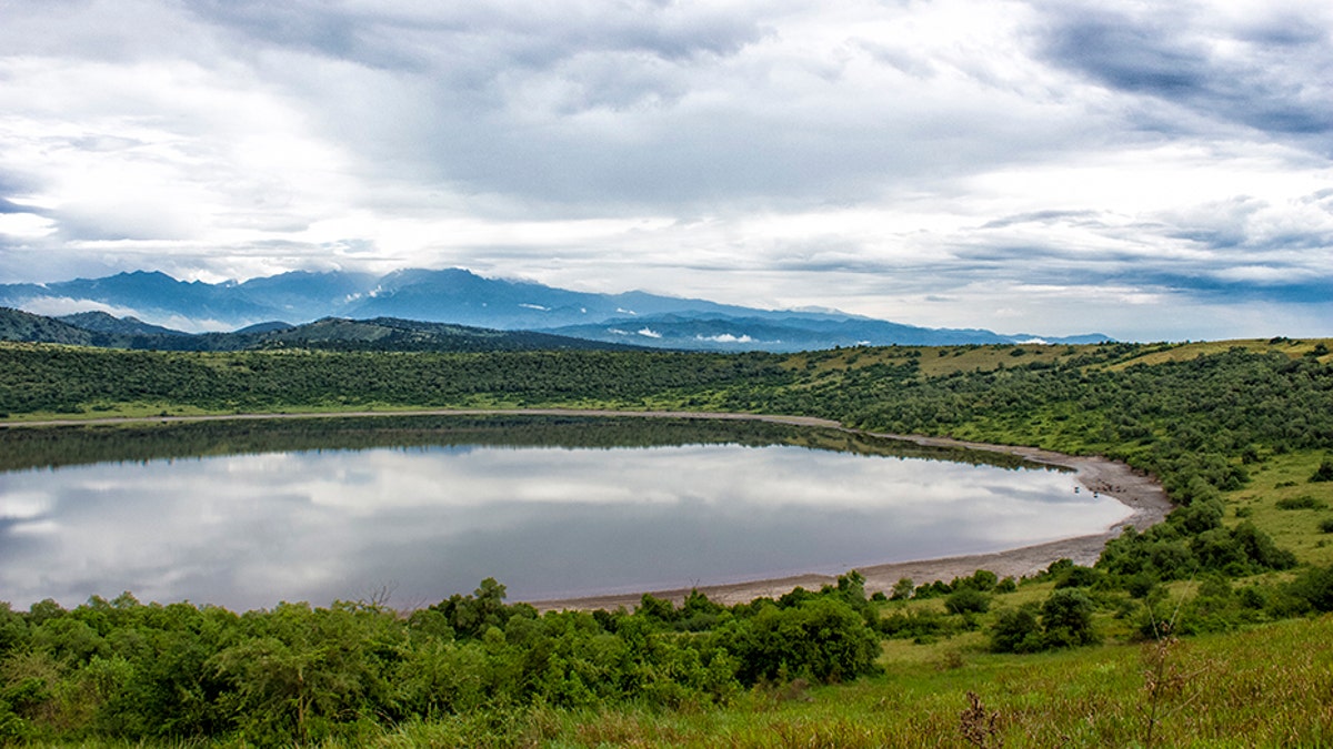 Moody clouds over a serene crater lake and blue mountains in Queen Elisabeth national park in Uganda. There are water buffalos grazing on the shore of the lake.