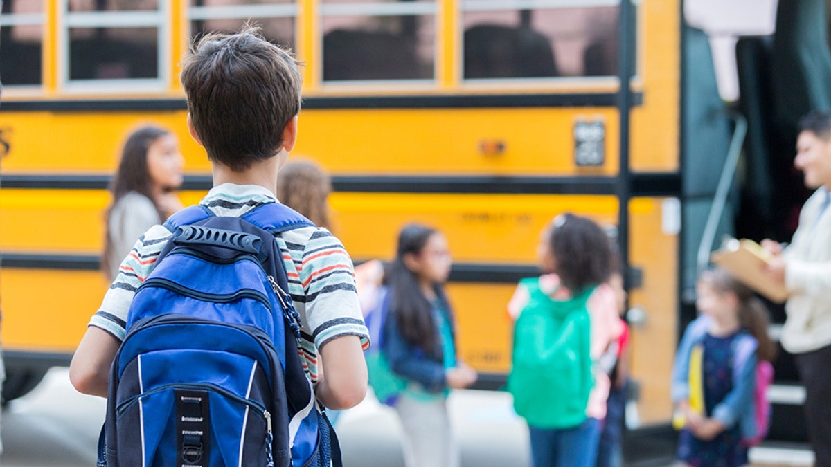 Rear view of elementary age boy waiting to get on school bus. His classmates are loading the bus in the background.