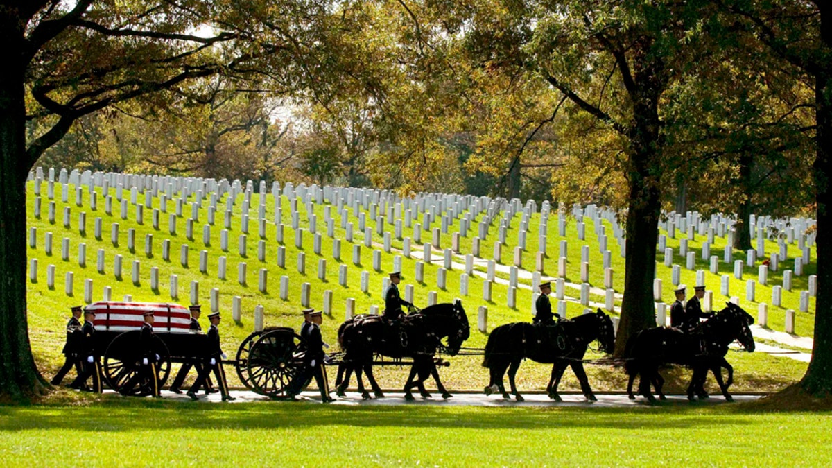 A flag-draped casket is carried by a horse-drawn caisson through Arlington National Cemetery during a full honors military funeral procession, with uniformed service members marching in solemn formation amid rows of headstones.