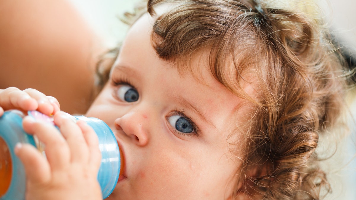 Close-up portrait of cute baby boy with blue eyes and brown curly hair drinking milk or juice from bottle.