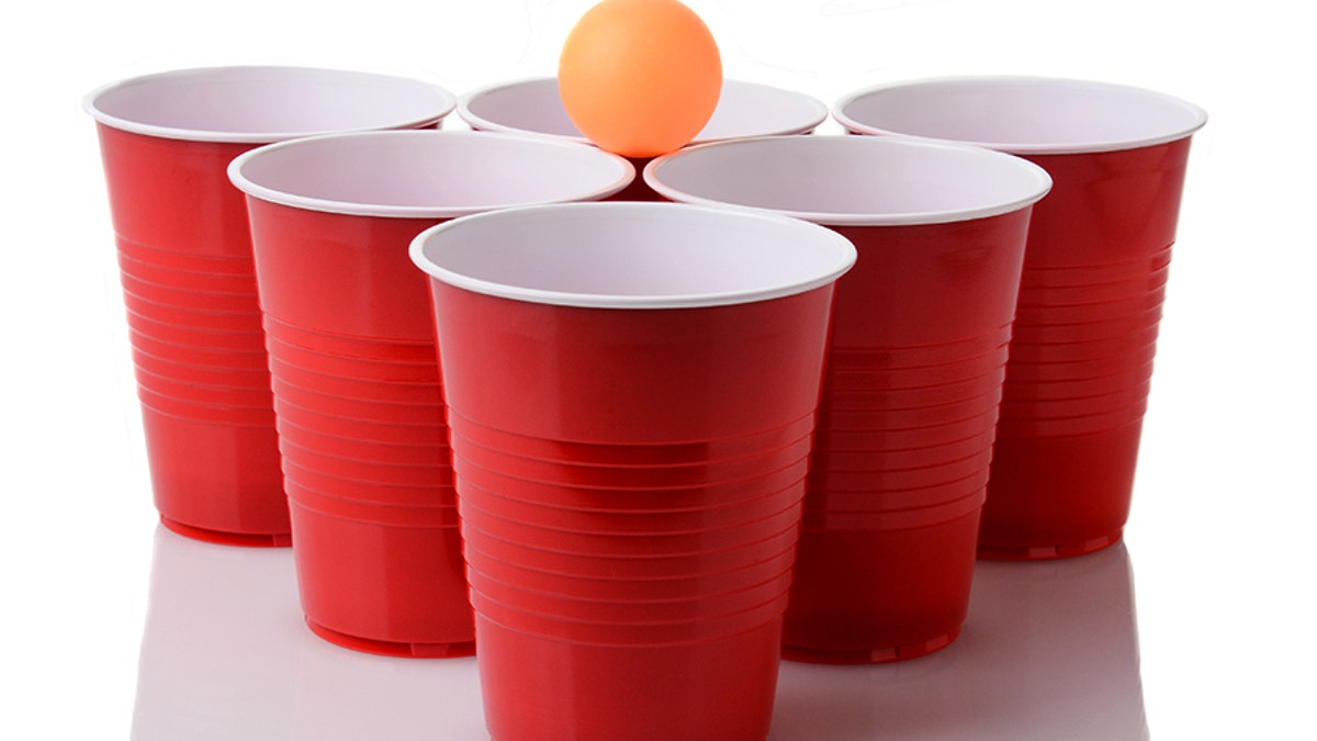 A yellow ping pong ball resting on a group of red plastic cups arranged for playing Beer Pong isolated on a white background with reflection.