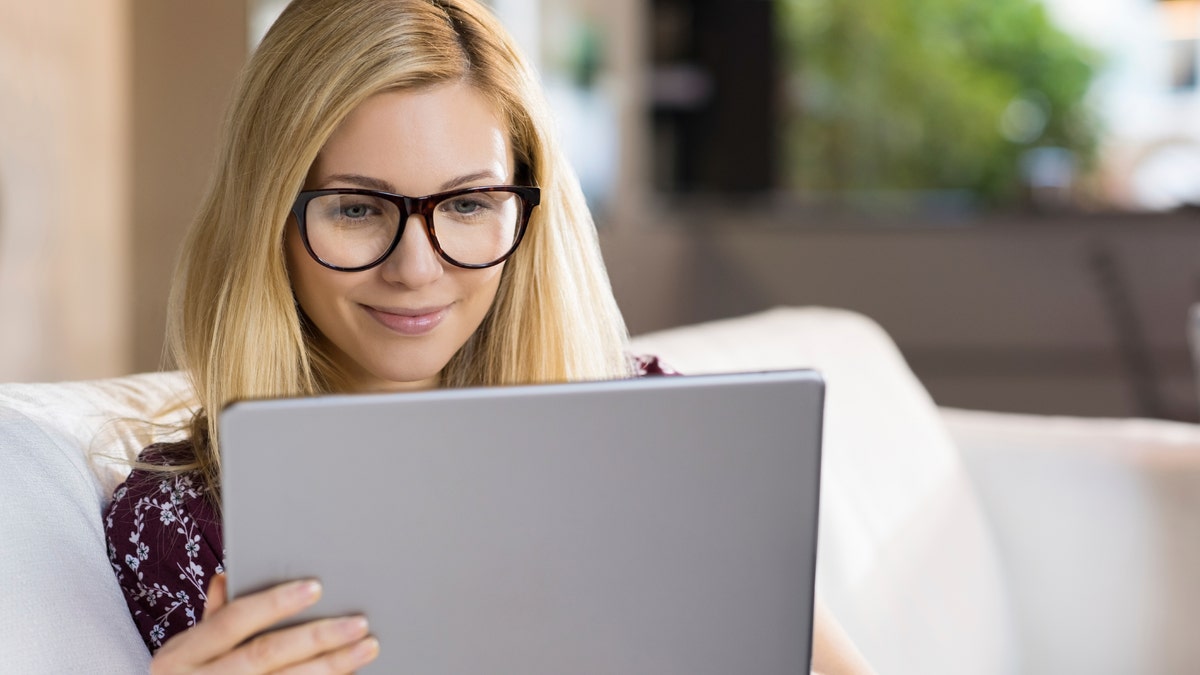 Happy young blonde woman sitting on sofa and using digital tablet. Happy young student with eyeglasses watching a movie on tablet. Female student studying on tablet at home.