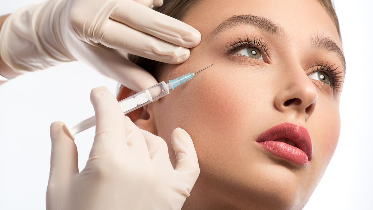 Serine young woman is getting facial botox injection. Beautician hands in gloves holding syringe near her face