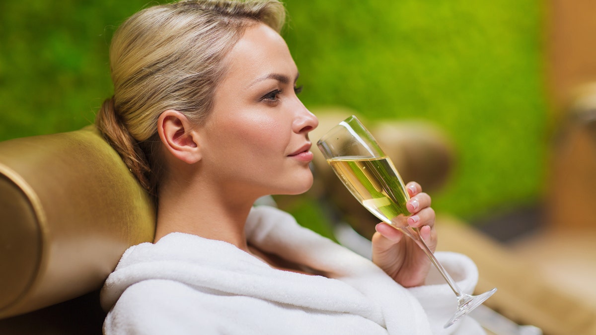 people, beauty, lifestyle, holidays and relaxation concept - beautiful young woman in white bath robe lying on chaise-longue and drinking champagne at spa