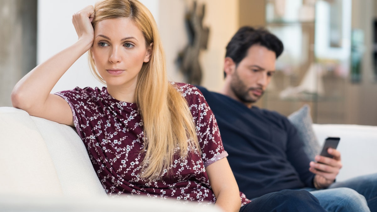 Young woman getting bored while man using phone in the background. Beautiful young woman feeling annoyed as man texting on phone. Young woman after an argument with her boyfriend in their living room.