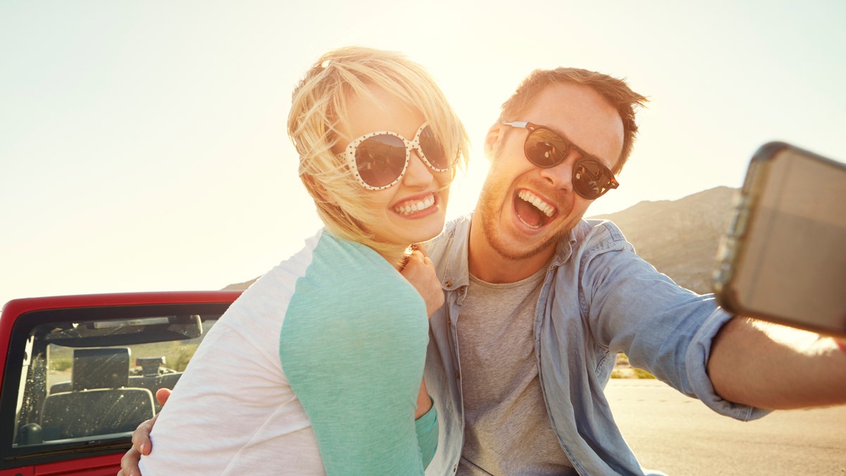 Couple On Road Trip Sit On Convertible Car Taking Selfie