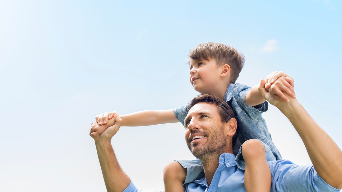 Father giving son ride on back in park. Portrait of happy father giving son piggyback ride on his shoulders and looking up. Cute boy with dad playing outdoor.