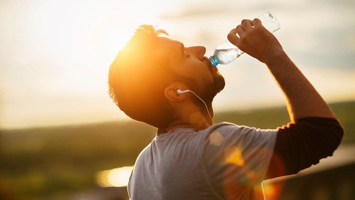 Close up of a young man drinking water from a plastic bottle underneath a clear sky. He is listening to music from small white earphones and his eyes are closed. The sun is shining strong in the background.