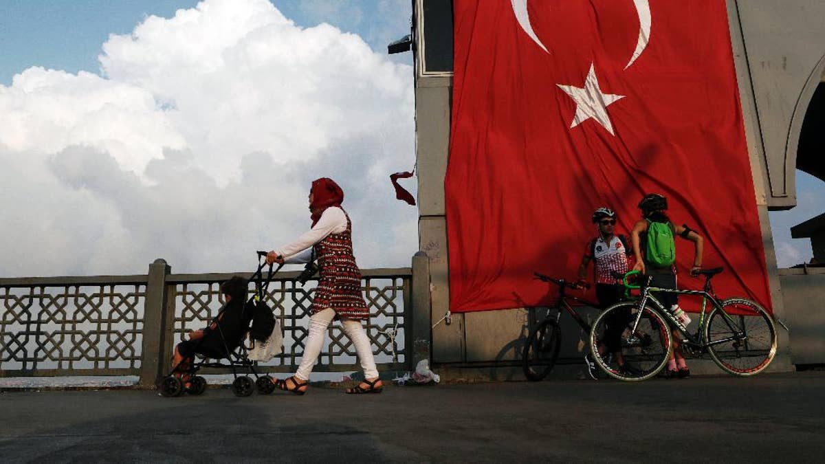 A woman passes with her child by two cyclist as they stand by a giant Turkey flag at the Galata bridge in Istanbul, on Thursday, Aug. 4, 2016. The Turkish government characterizes the movement of Fethullah Gulen, who lives in self-imposed exile in Pennsylvania, as a terrorist organization. (AP Photo/Petros Karadjias)
