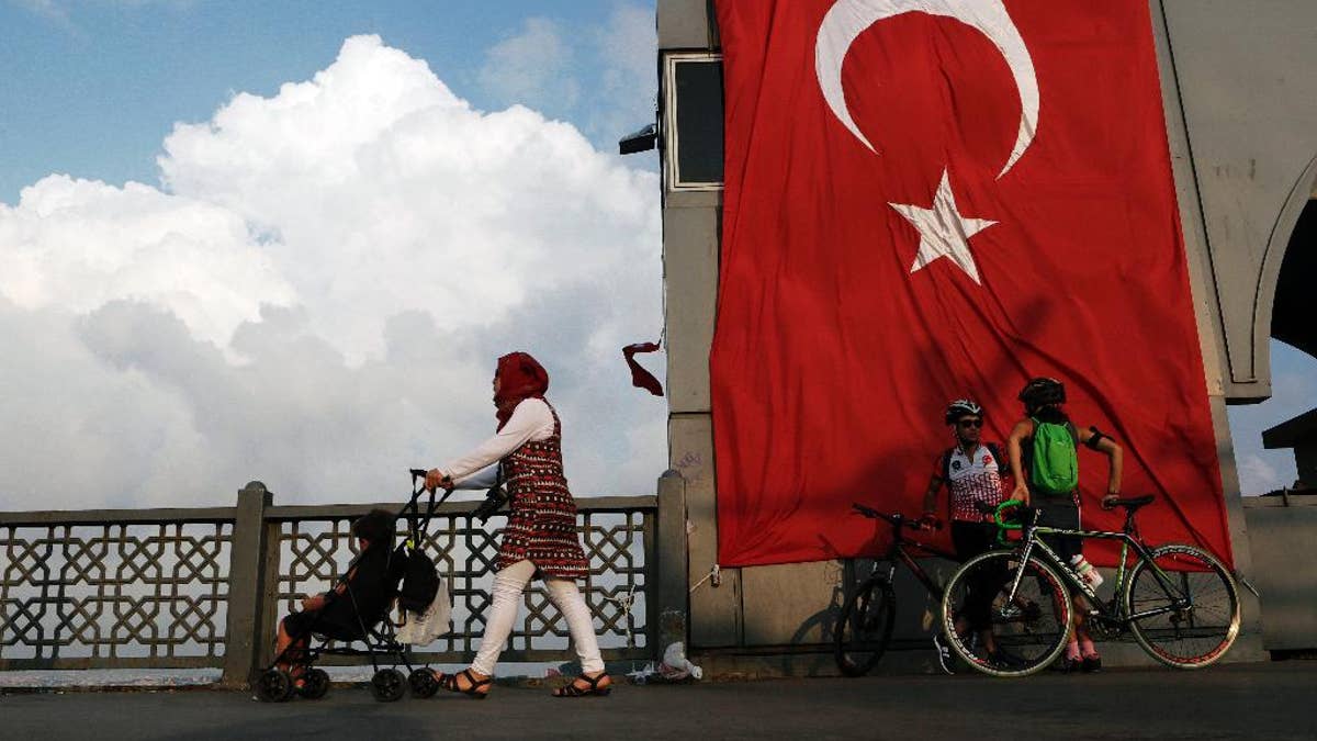 A woman passes with her child by two cyclist as they stand by a giant Turkey flag at the Galata bridge in Istanbul, on Thursday, Aug. 4, 2016. The Turkish government characterizes the movement of Fethullah Gulen, who lives in self-imposed exile in Pennsylvania, as a terrorist organization. (AP Photo/Petros Karadjias)