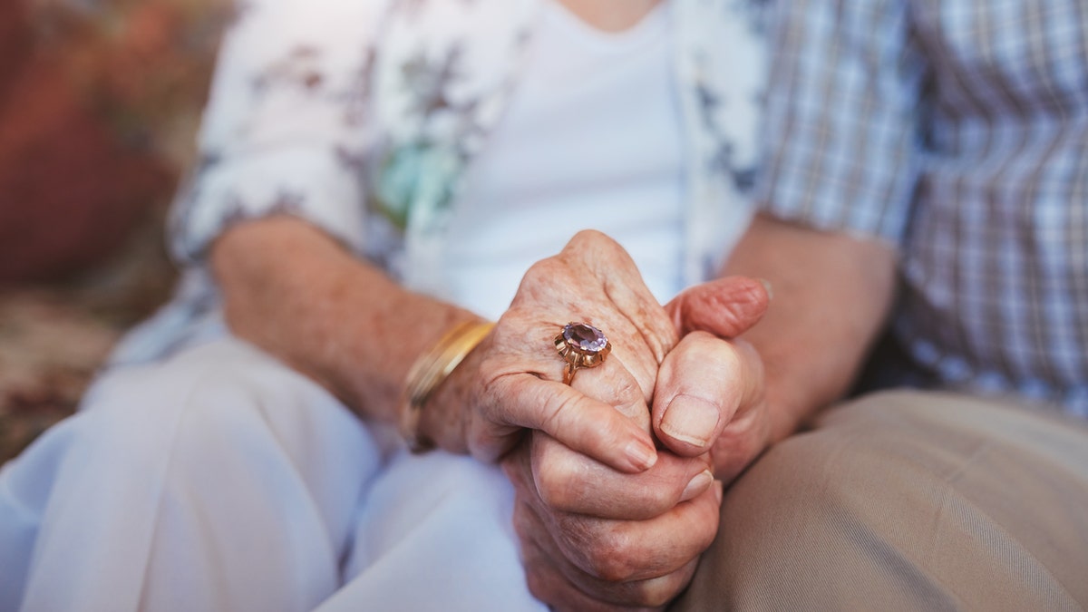 Cropped shot of elderly couple holding hands while sitting together at home. Focus on hands.