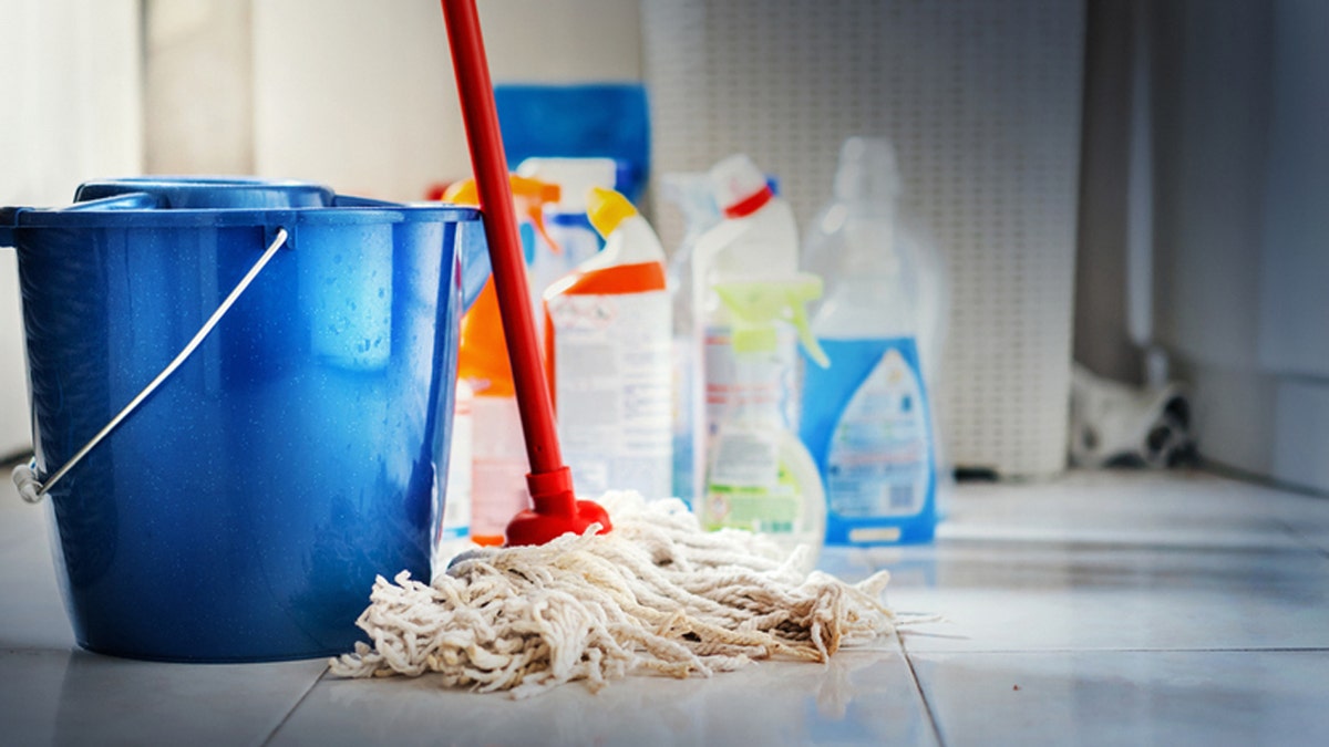 Closeup of home cleaning products with blue bucket and a mop in front in sharp focus. All products placed on white and poorly lit bathroom floor.