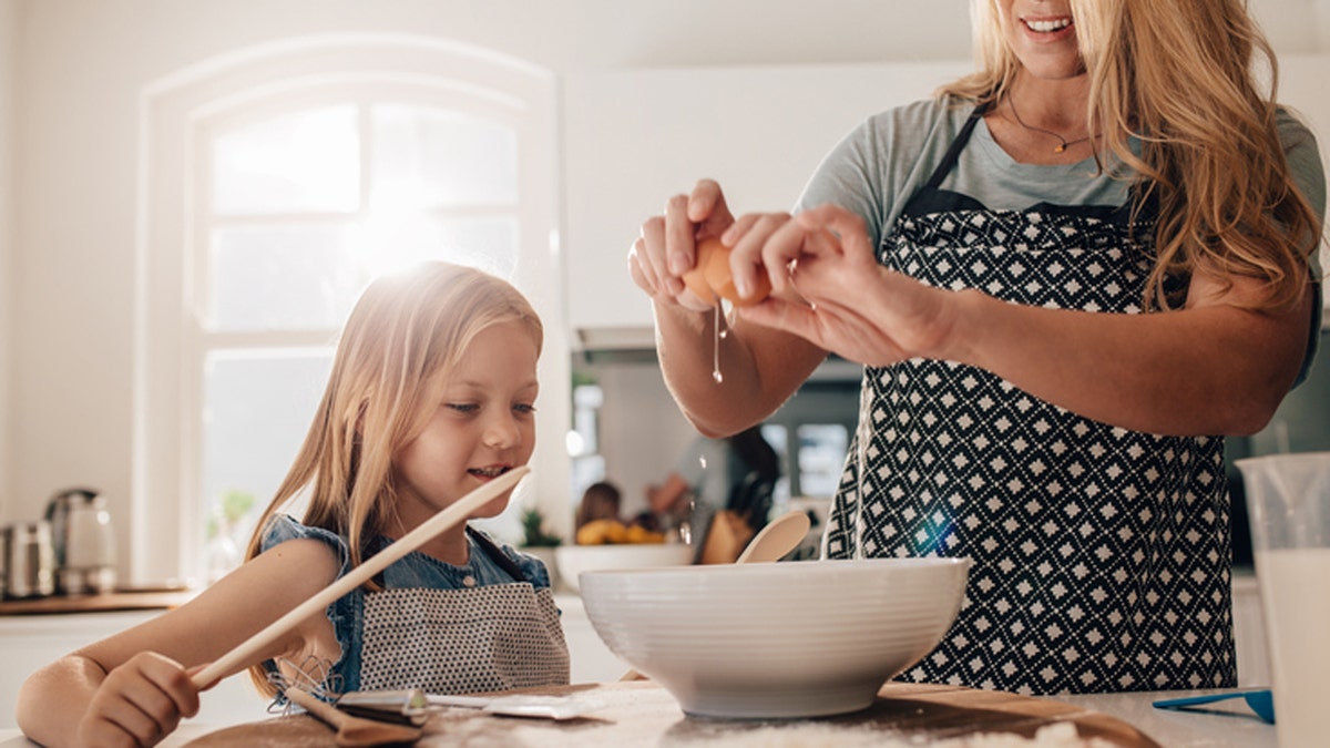 Mother cracking an egg into bowl with her daughter in kitchen. Woman and little girl preparing food in kitchen.