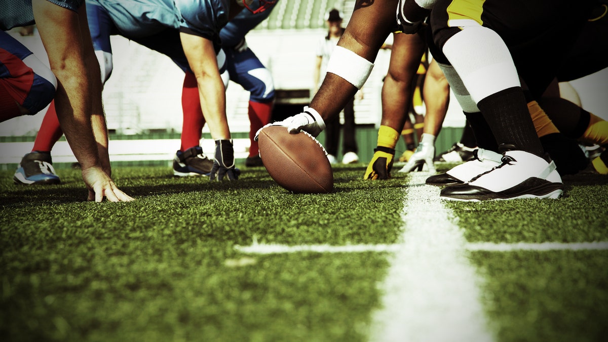 The blue, red, and white football team line up at the line of scrimmage against the black and yellow team. The play is about to begin as the center has hand on the football and is ready to snap to the quarterback. Photo perspective is low and between the two teams.