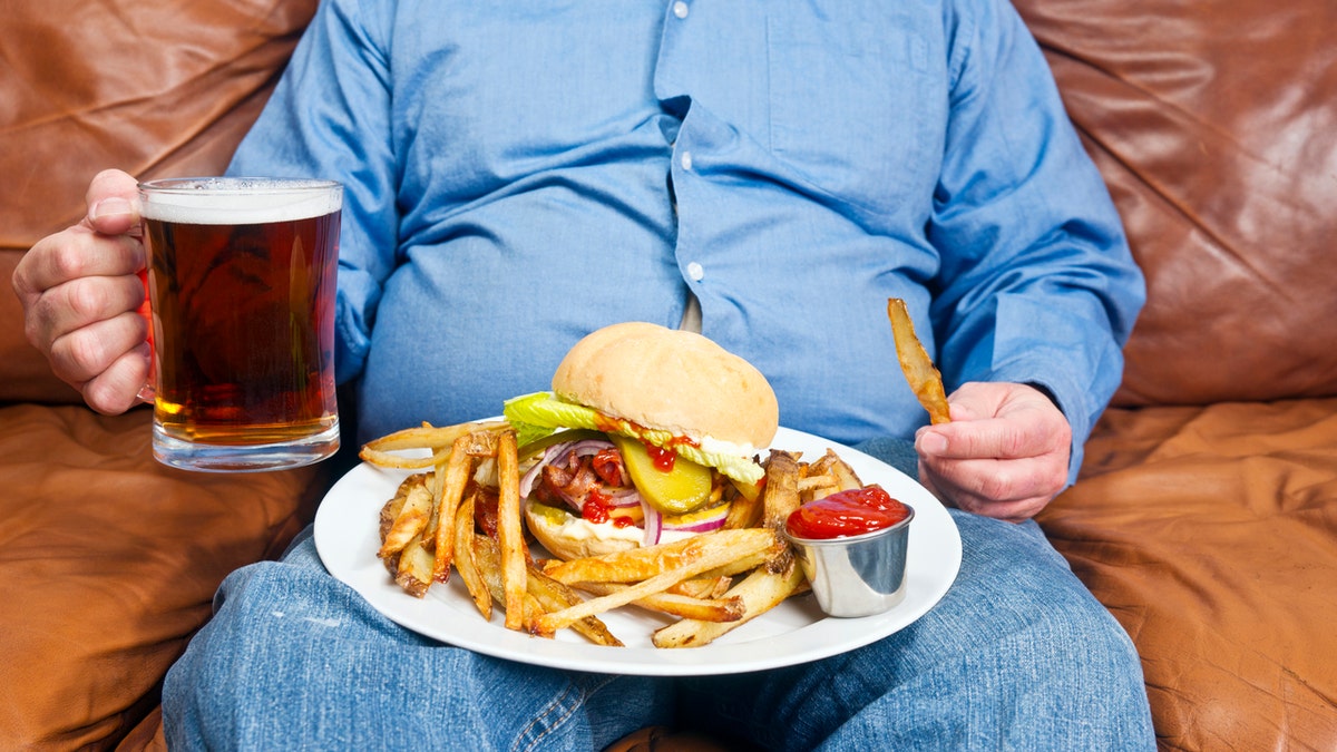 A photo of an overweight man sitting on an old couch with a very large unhealthy meal on his lap and a pint of beer in his hand. Obesity is a major cause of diabetes.